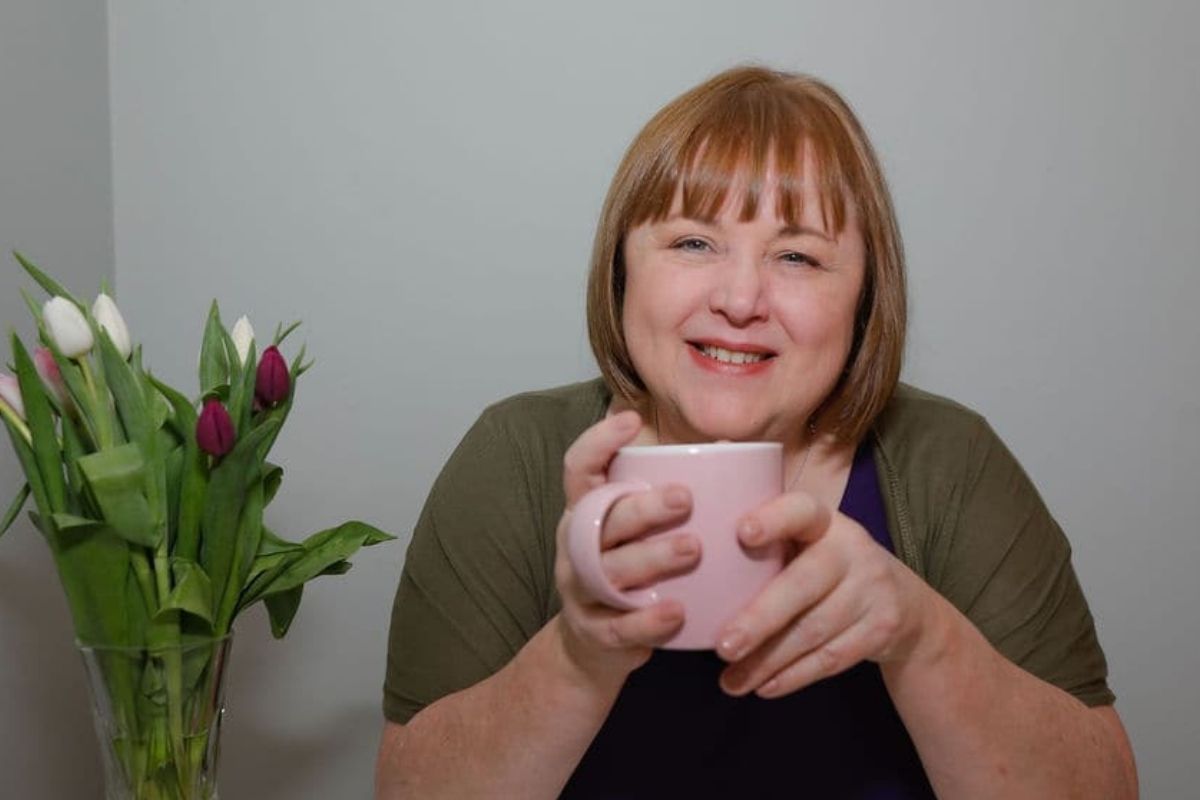 Jackie McGloughlin holistic therapist at Energy Flows welcomes you here in Maynooth and online for Jin Shin Jyutsu, Reflexology, and Reiki treatments. She is smiling while holding a pink mug, at a white table with tulips on the left.